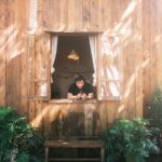 A person relaxes by the window of a rustic wooden cabin, surrounded by lush greenery in bright summer sunlight.
