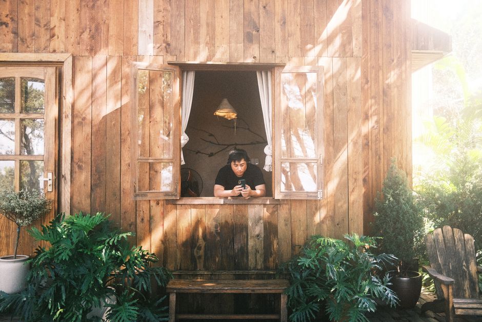 A person relaxes by the window of a rustic wooden cabin, surrounded by lush greenery in bright summer sunlight.
