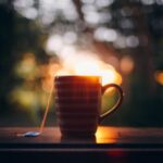 Ceramic mug with teabag placed on wooden table against blurred green park during sunset