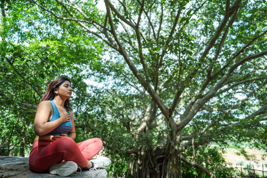 A woman meditates in a serene outdoor setting, surrounded by lush greenery.