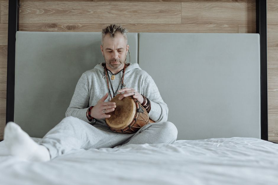 Relaxed man playing a djembe drum on a bed in a cozy, indoor setting.