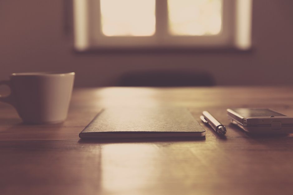 A minimalist workspace setup featuring a cup, notebook, pen, and smartphone on a wooden desk indoors.