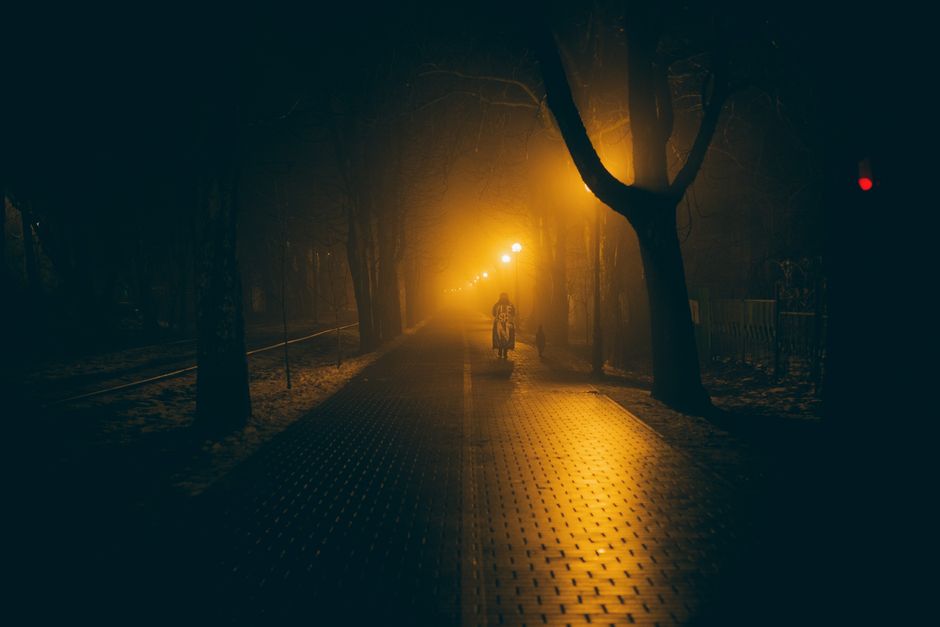 A solitary figure walking on a foggy street lit by orange streetlights at night.