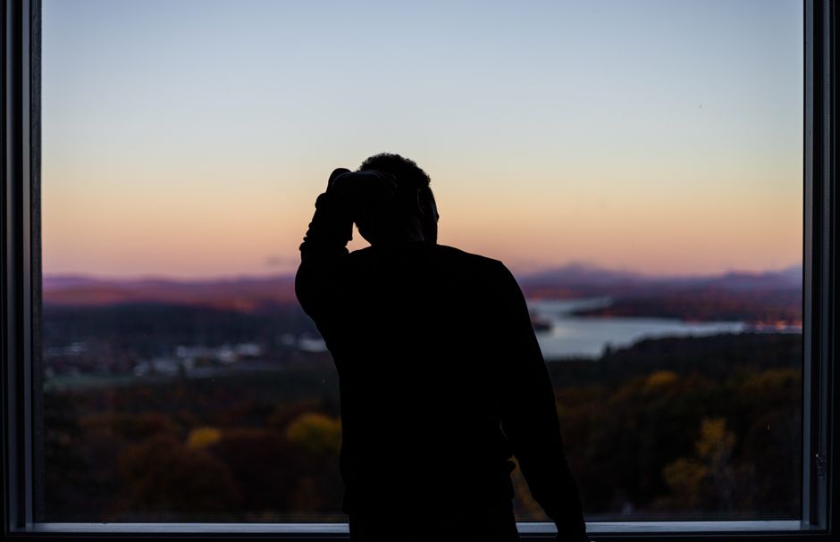 A silhouette of a person at a window, gazing at a scenic lake and mountain landscape during sunset.